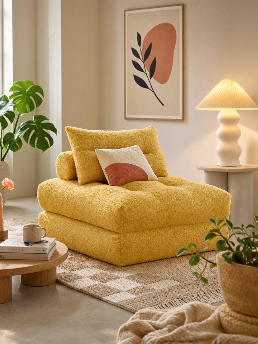 A mustard-yellow bouclé sofa bed with a round-patterned cushion, in a sunlit room beside a pleated lamp and a monstera plant
